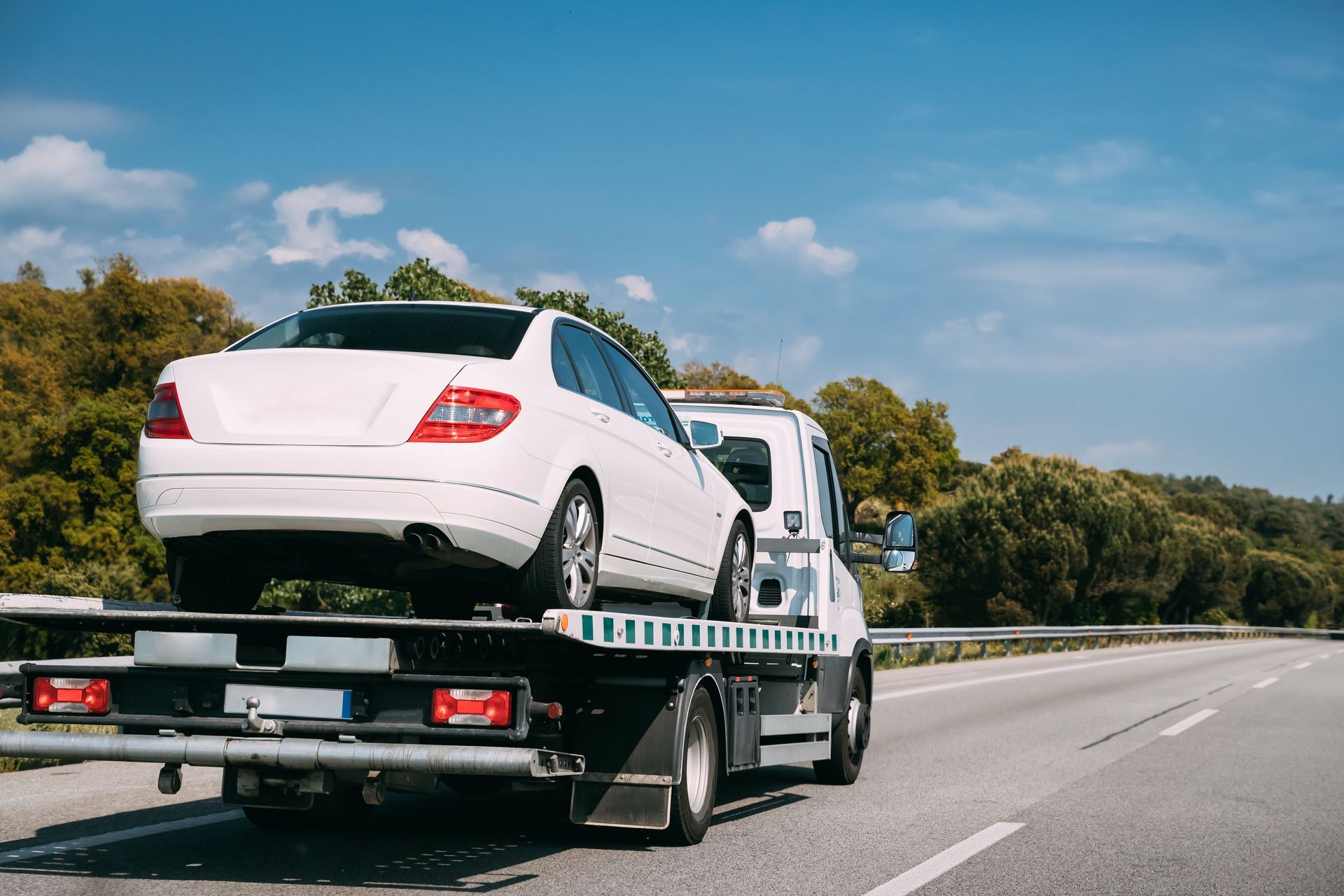 White car on a tow truck, roadside setting with trees and a blue sky.