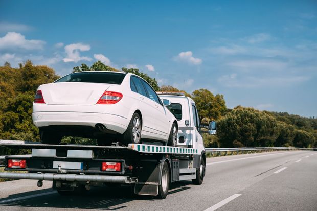 White car on a tow truck, roadside setting with trees and a blue sky.