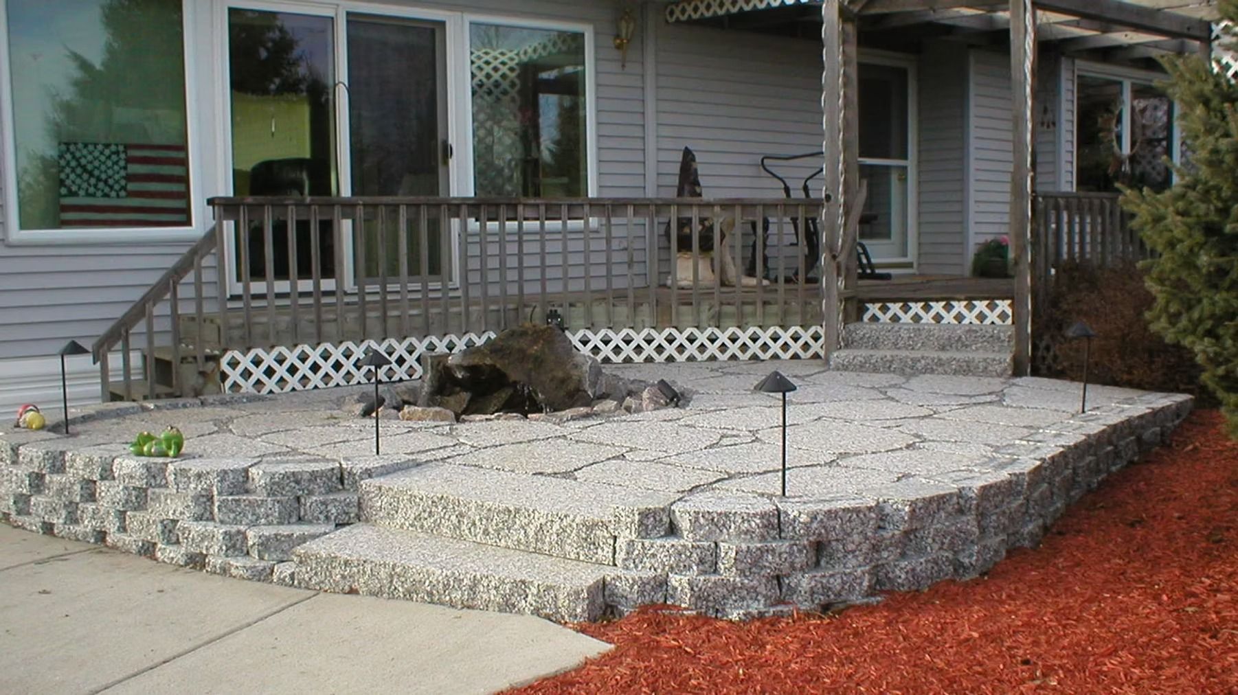 Front porch with wooden railing and stone landscaping, bordered by a concrete walkway and red mulch.