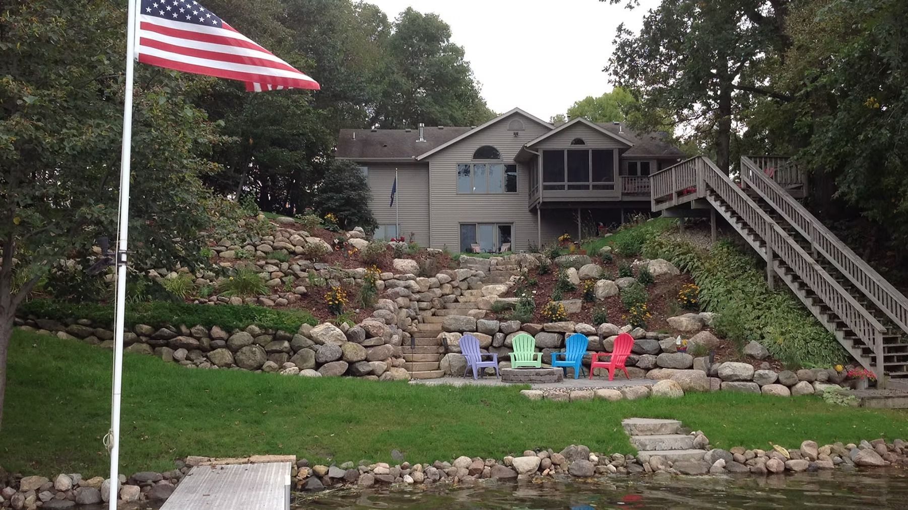 House on a hillside with an American flag, stone retaining wall, and colorful chairs on the lawn