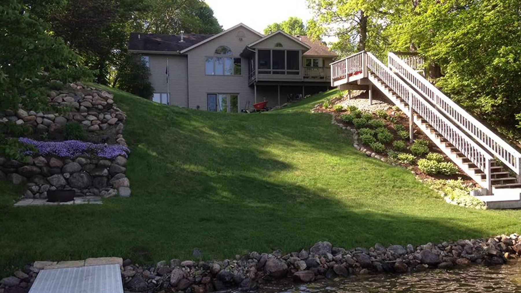 Backyard hillside with stone retaining wall, green lawn, and a house with a wooden deck and stairs