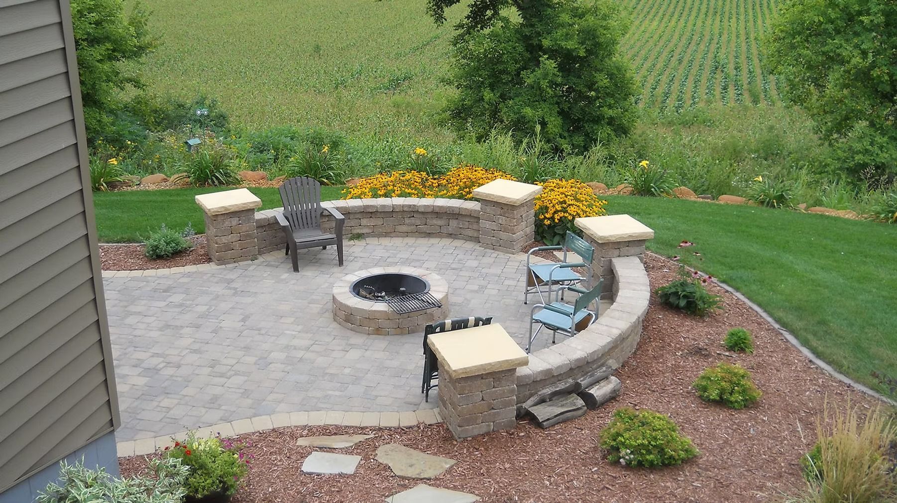 Backyard patio with stone fire pit, curved benches, and flower beds beside a grassy lawn