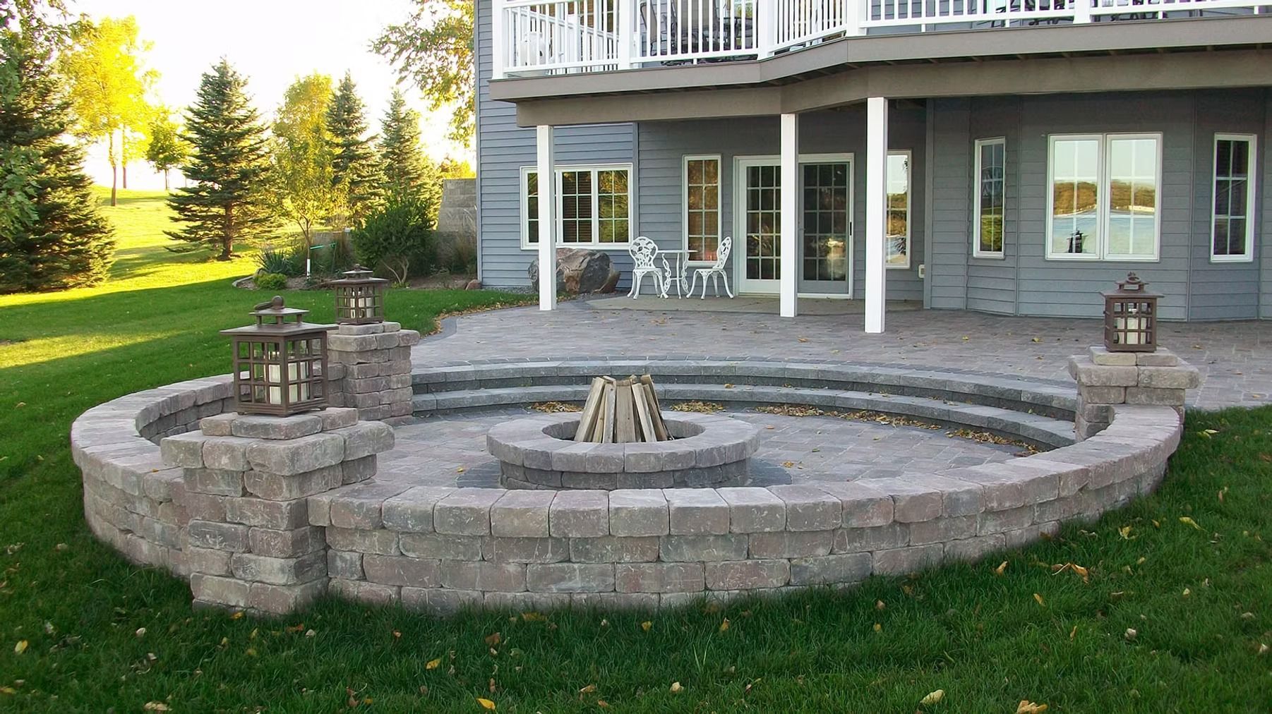 Stone fire pit on a curved patio in a green backyard beside a house with covered porch
