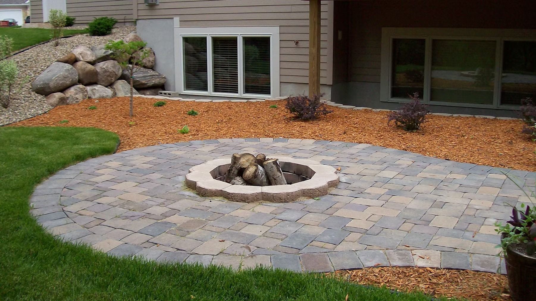 Stone patio with circular fire pit, bordered by mulch, grass, and a house exterior