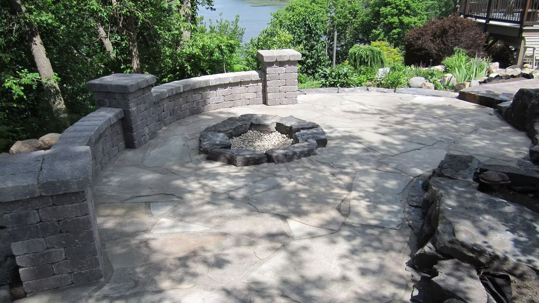 Stone patio with a circular fire pit, low stone walls, and surrounding greenery in a landscaped backyard