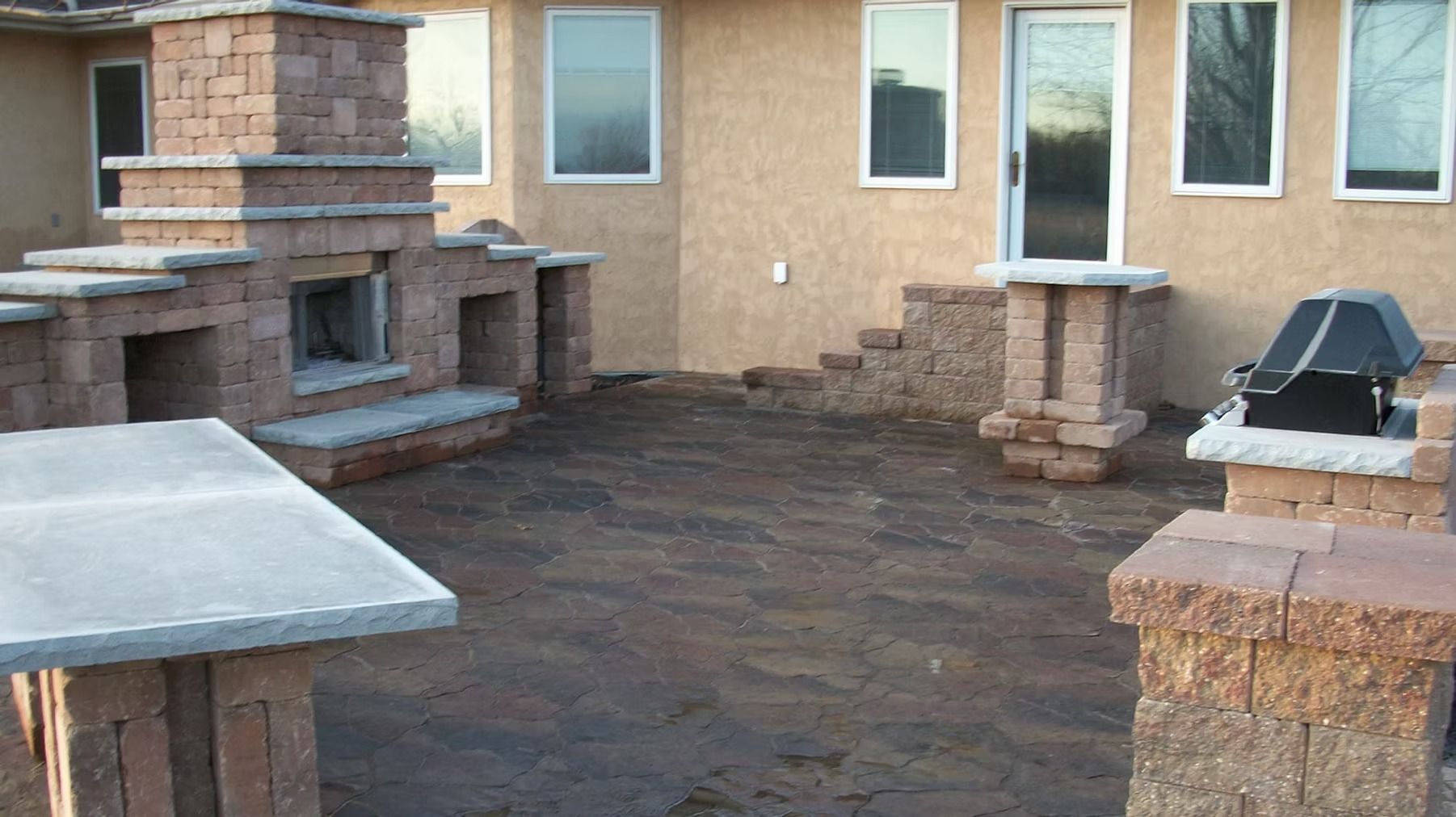 Stone patio with built-in outdoor grill and benches beside a beige house