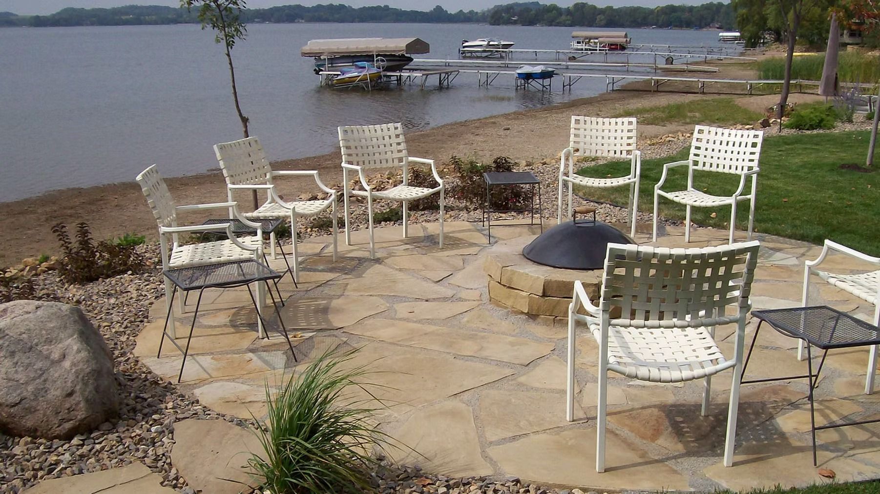Patio chairs around a fire pit by a lakeshore with boats docked in the background