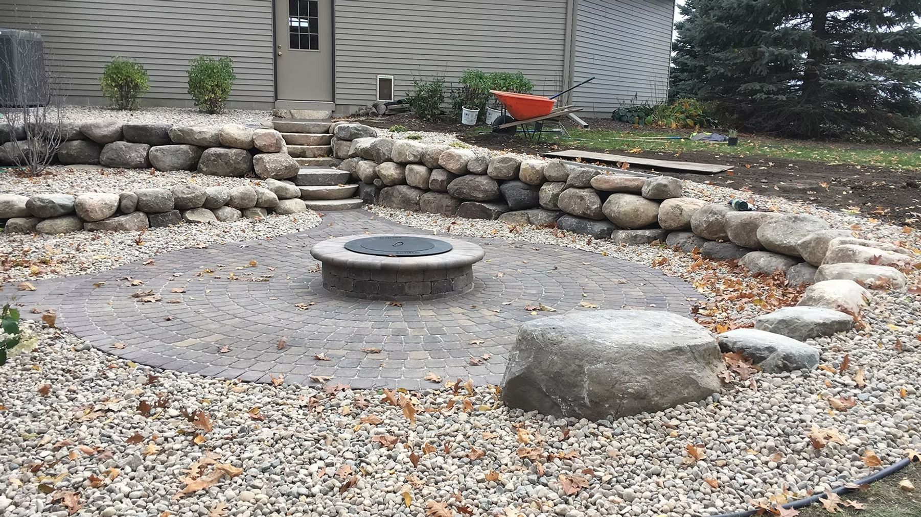 Rock-ringed backyard fire pit with gravel ground and a central metal bowl near a house.