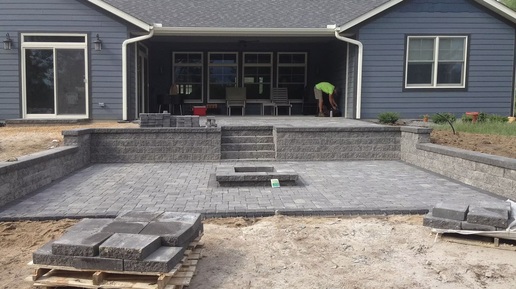 Backyard patio under construction beside a blue house, with pavers and a worker in green near the deck