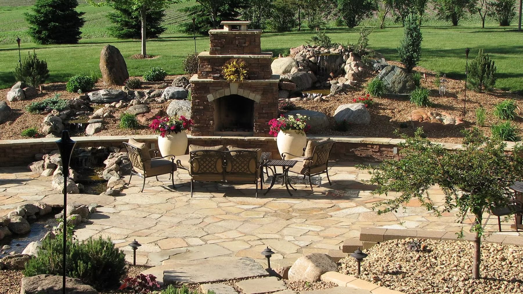 Stone backyard patio with built-in fireplace, outdoor seating, and landscaped garden beds