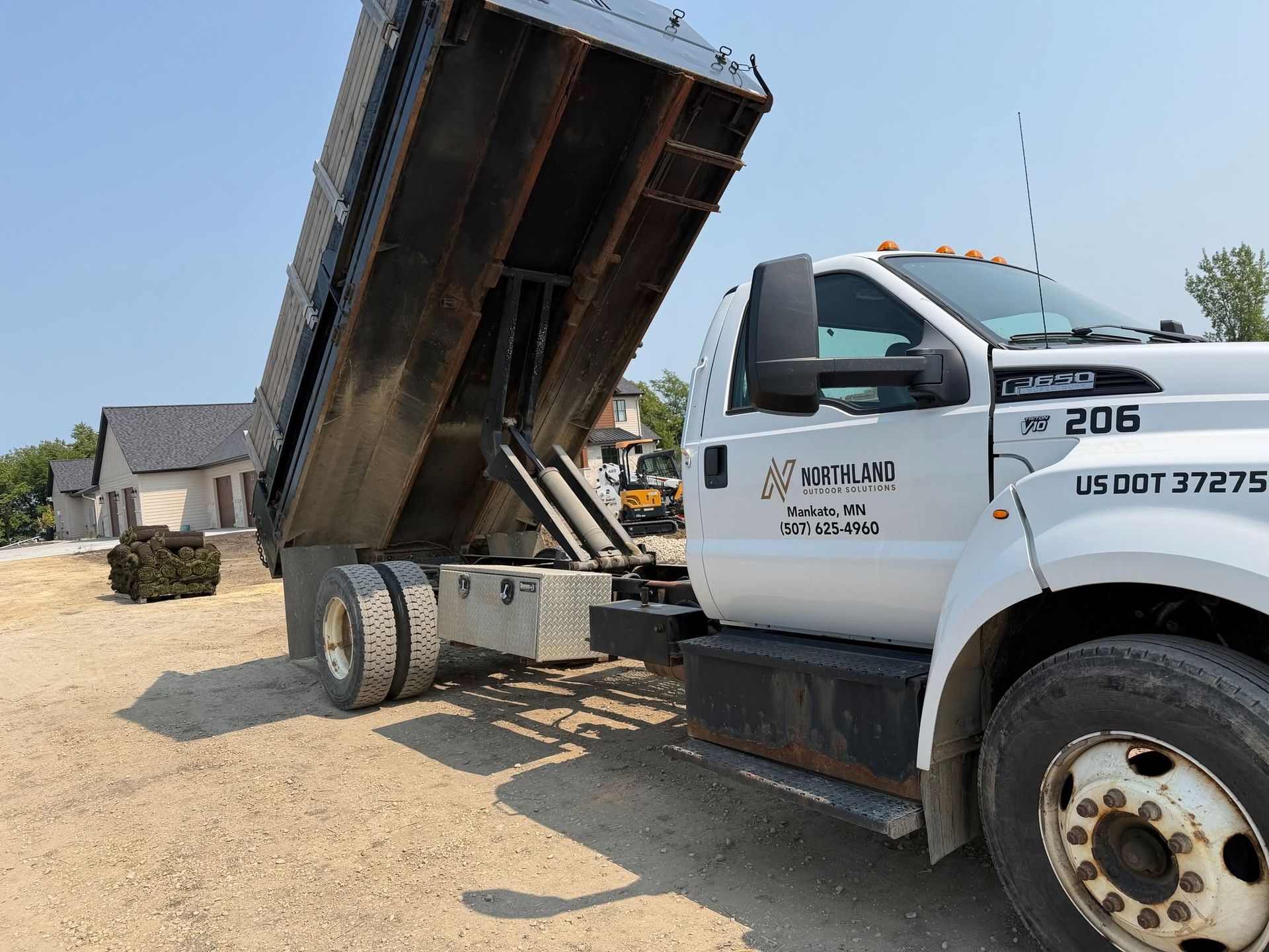 White dump truck with raised bed unloading on a dirt roadside near houses under a clear sky
