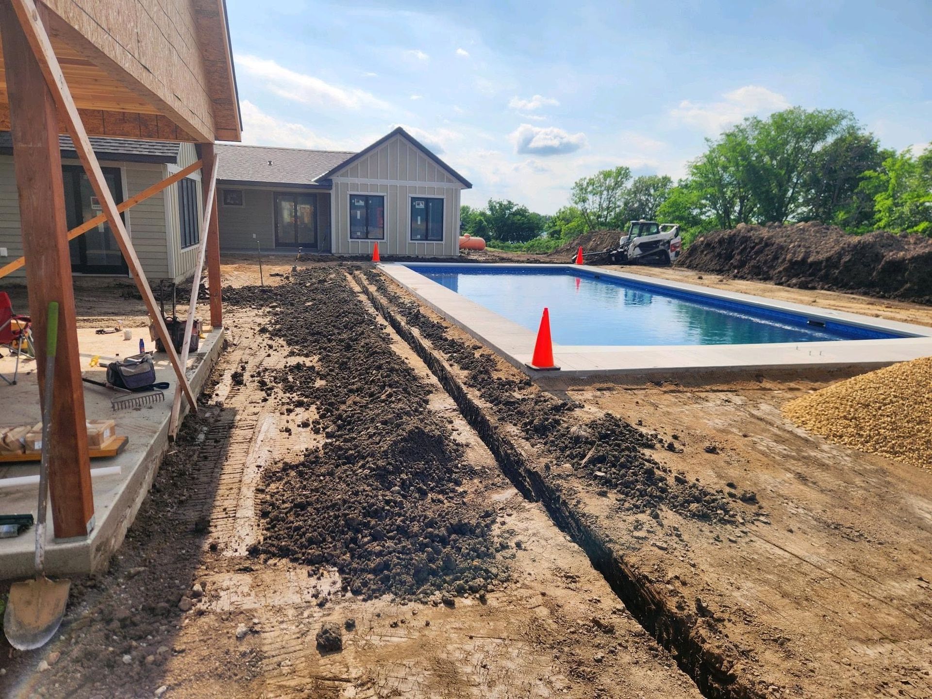 Backyard pool under construction beside a house, with concrete, dirt, and orange safety cones