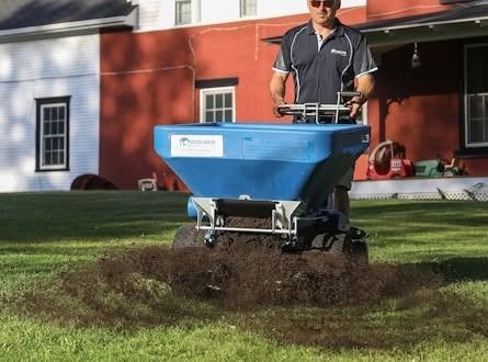 Man spreading fertilizer with a blue lawn spreader on a grassy yard in front of a red house
