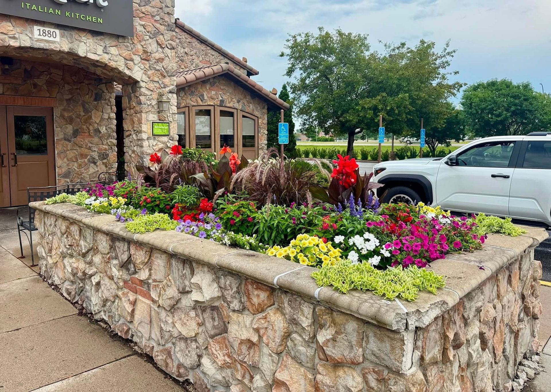 Stone planter with colorful flowers outside a rustic building, with parked cars nearby