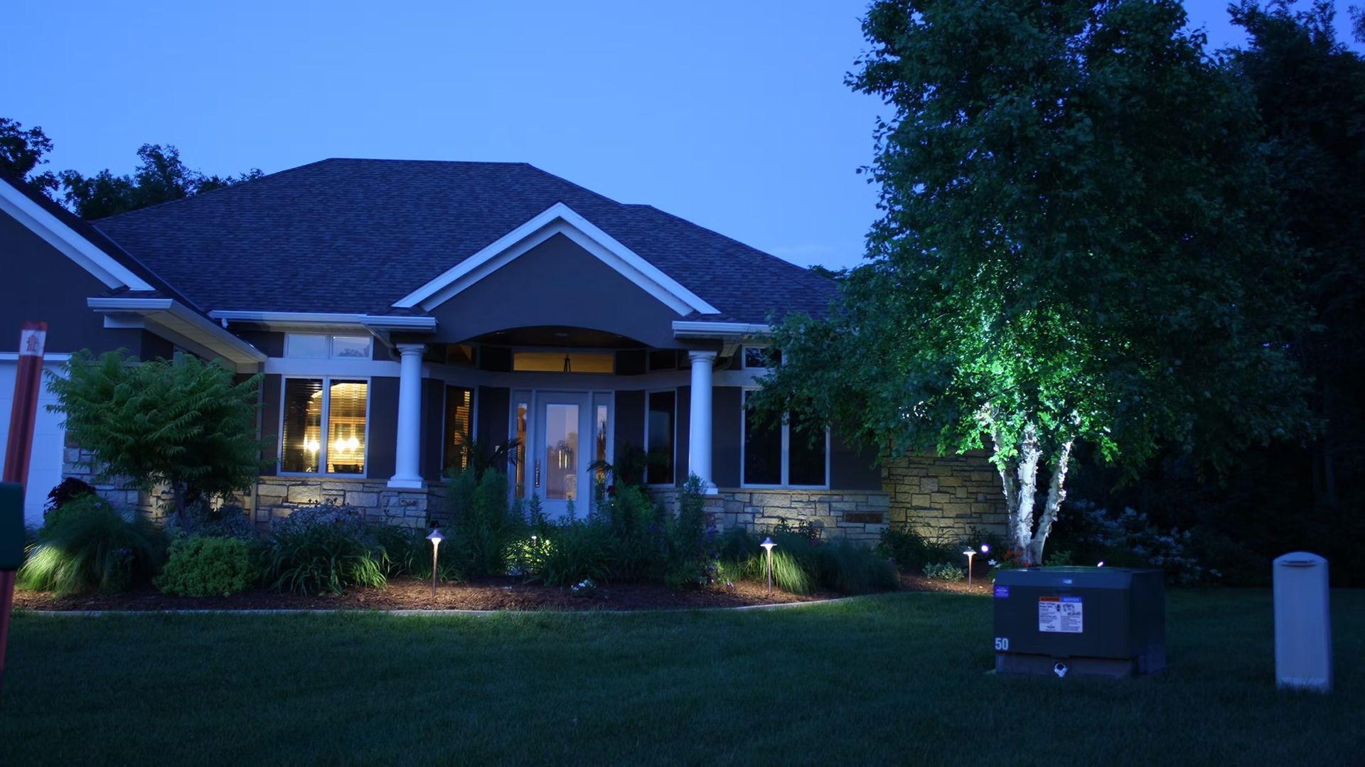 House with lit windows at dusk, front yard and trees illuminated by outdoor lights.