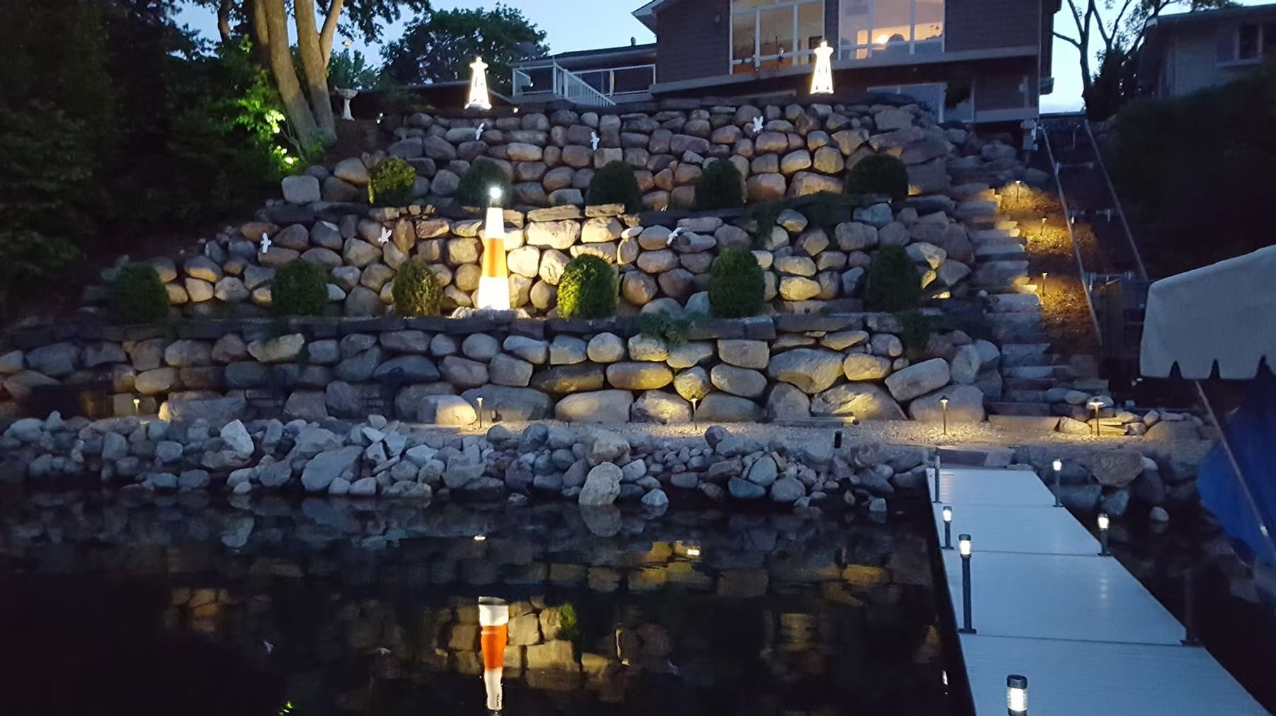 Stone terraced garden wall lit at dusk, reflected in a calm pond with a small house above.