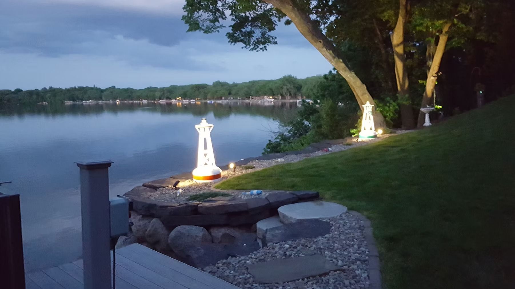 Lakeside dusk view with glowing garden lights, stone fire pit, and trees along the shore