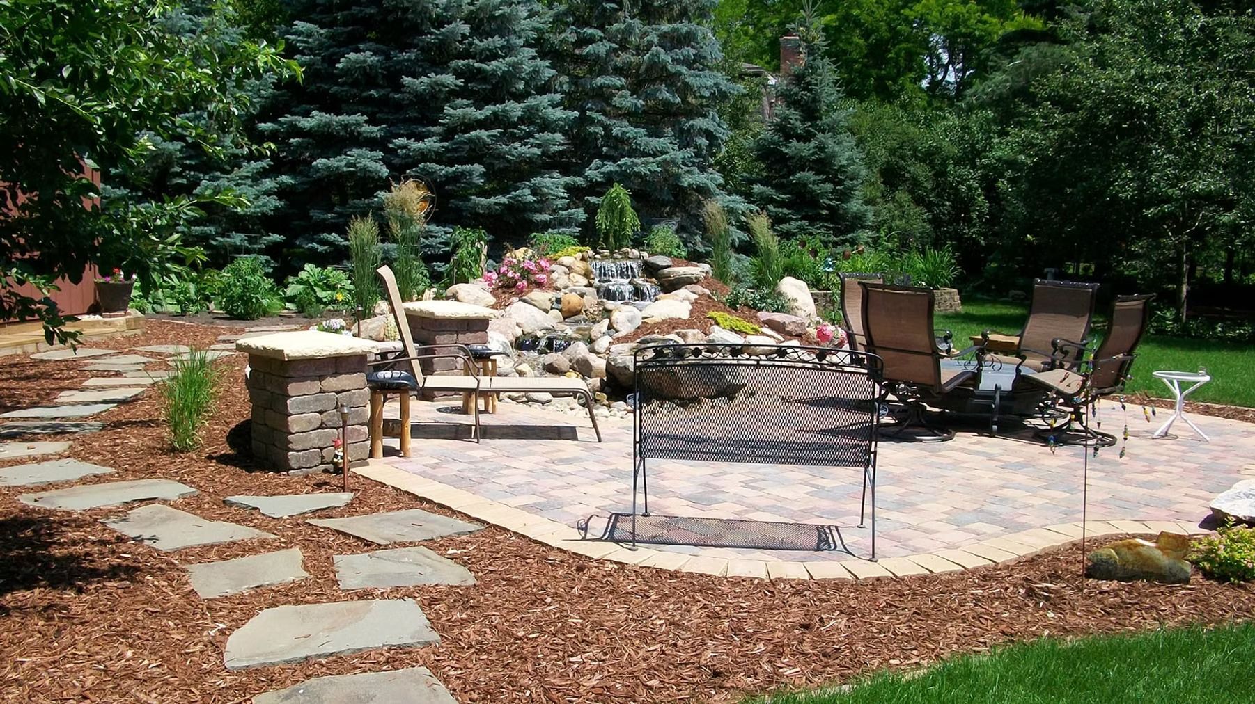 Stone patio with outdoor seating, fire pit, and landscaped garden surrounded by trees