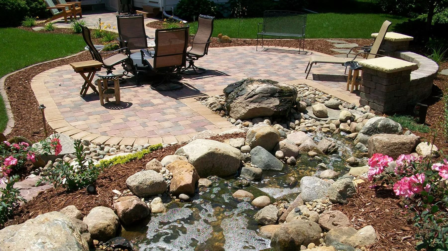 Stone patio with outdoor chairs and a round table beside a small garden stream and flowers