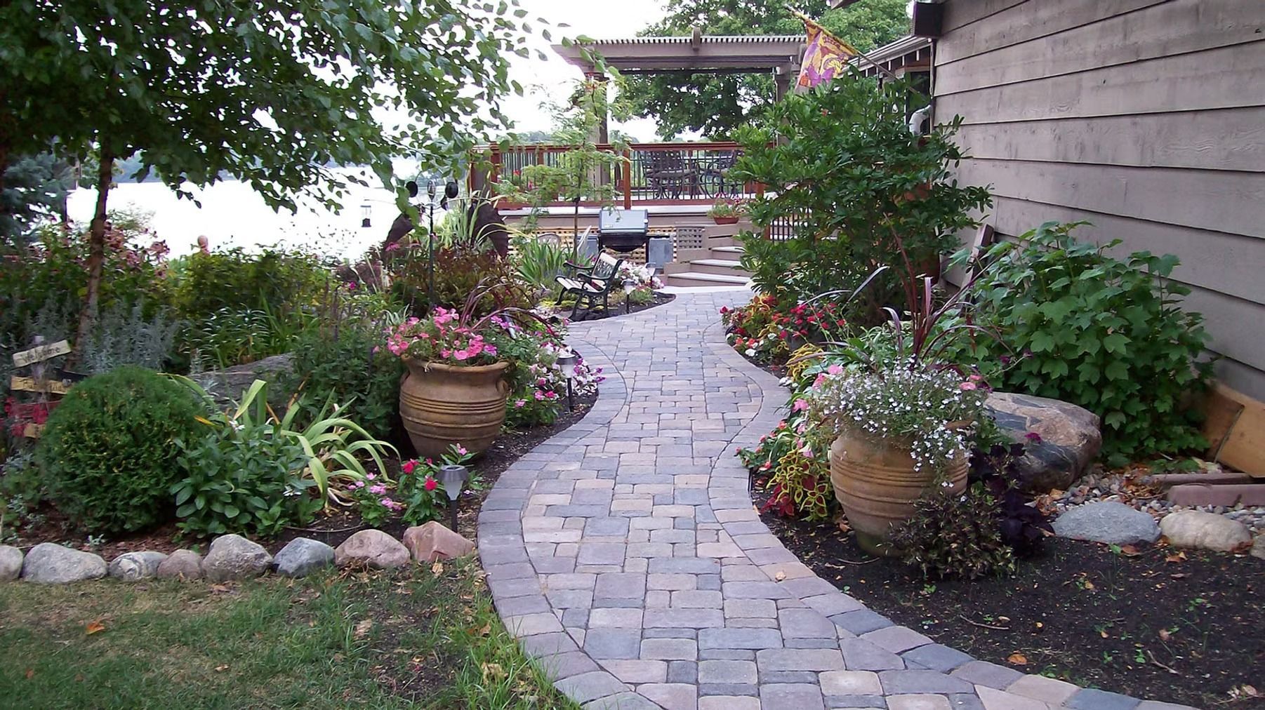 Curved stone path through a lush garden with potted flowers and greenery, leading to a wooden gate.