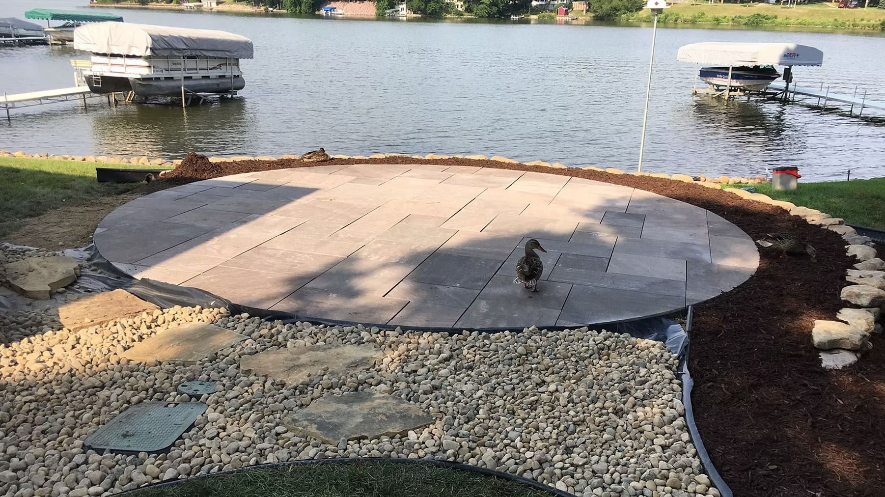 Circular concrete patio by a lakeside, bordered with gravel and mulch, with a duck near the center.
