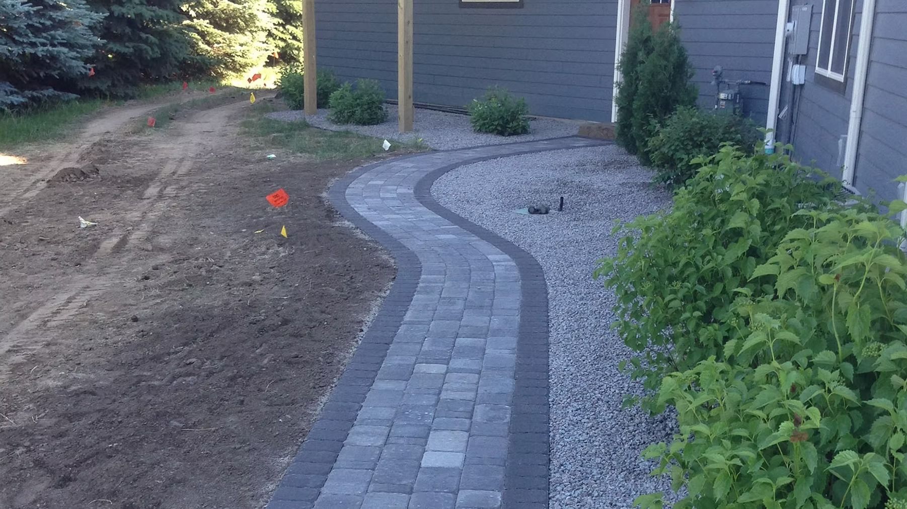 Curved brick walkway beside a house, bordered by gravel and plants, with a small red marker nearby.