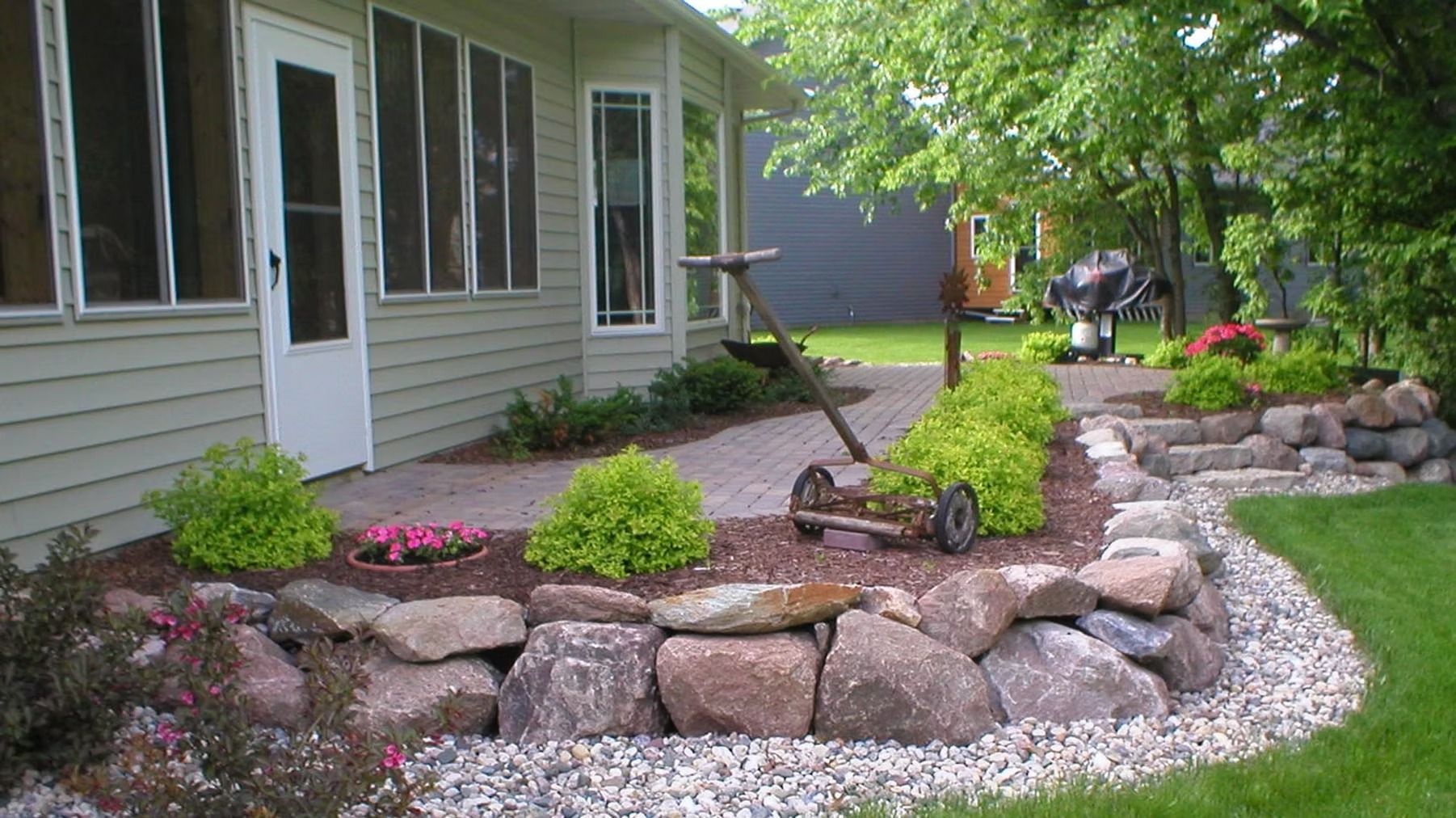 Landscaped yard with stone retaining walls, shrubs, and a house patio beside a gravel path