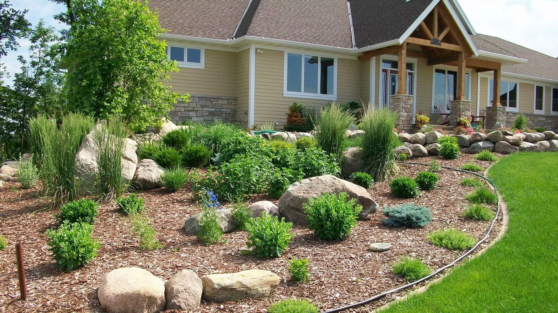 Landscaped front yard with rocks, shrubs, mulch, and a tan house with a covered porch