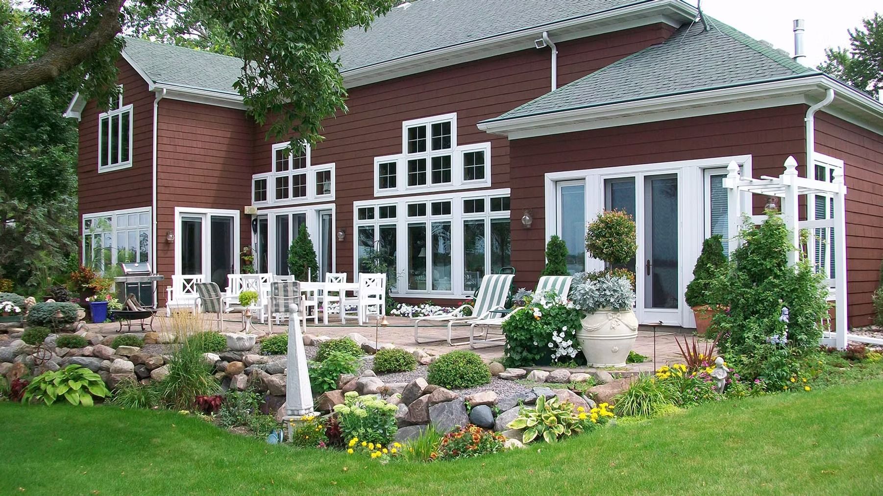 Red brick house with white trim, large windows, patio seating, and a lush flower-filled garden.