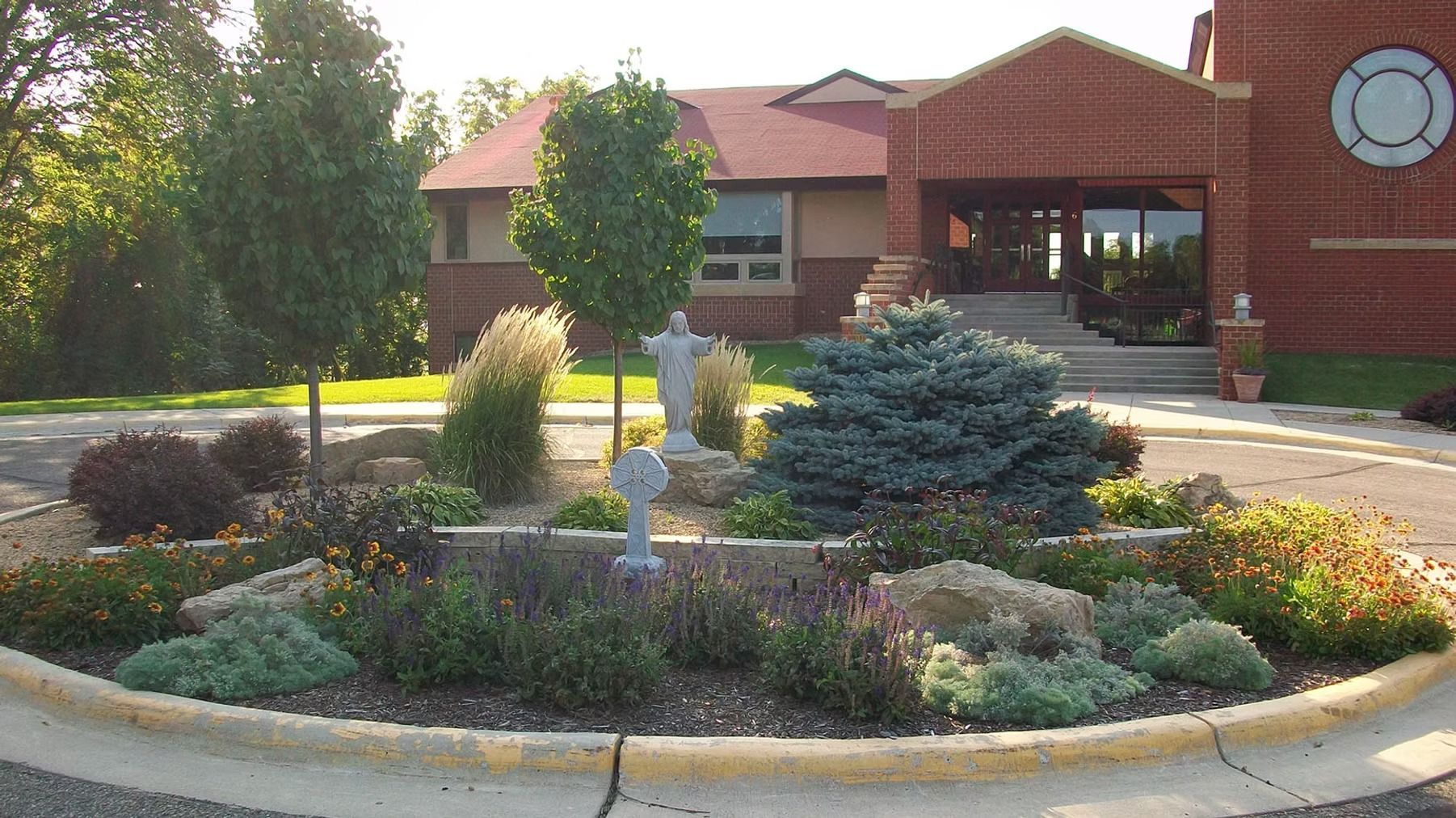Landscaped roundabout with flowers and shrubs in front of a brick building with a circular window.