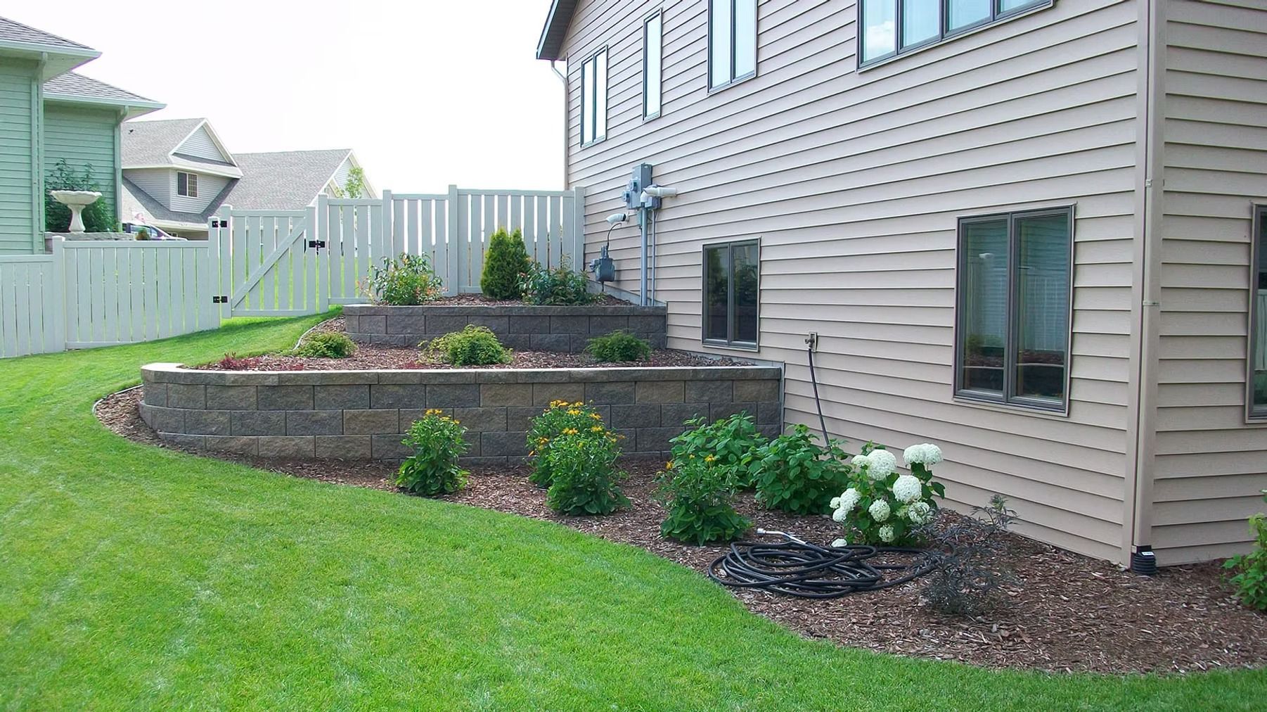 Backyard landscaping with tiered stone garden beds, shrubs, and green lawn beside a house