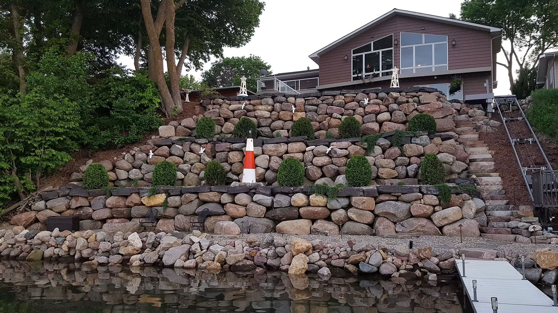 Stone retaining wall on a lakeshore beneath a house, with stairs and an orange marker near the water.