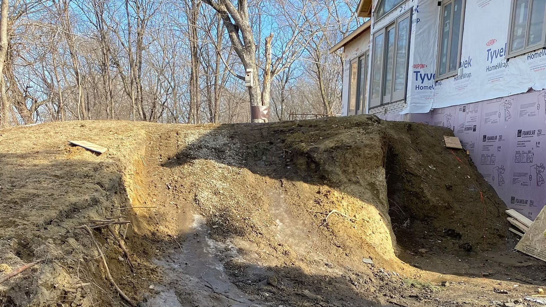 Mounded dirt and a steep excavation beside a house under construction, with bare trees in the background