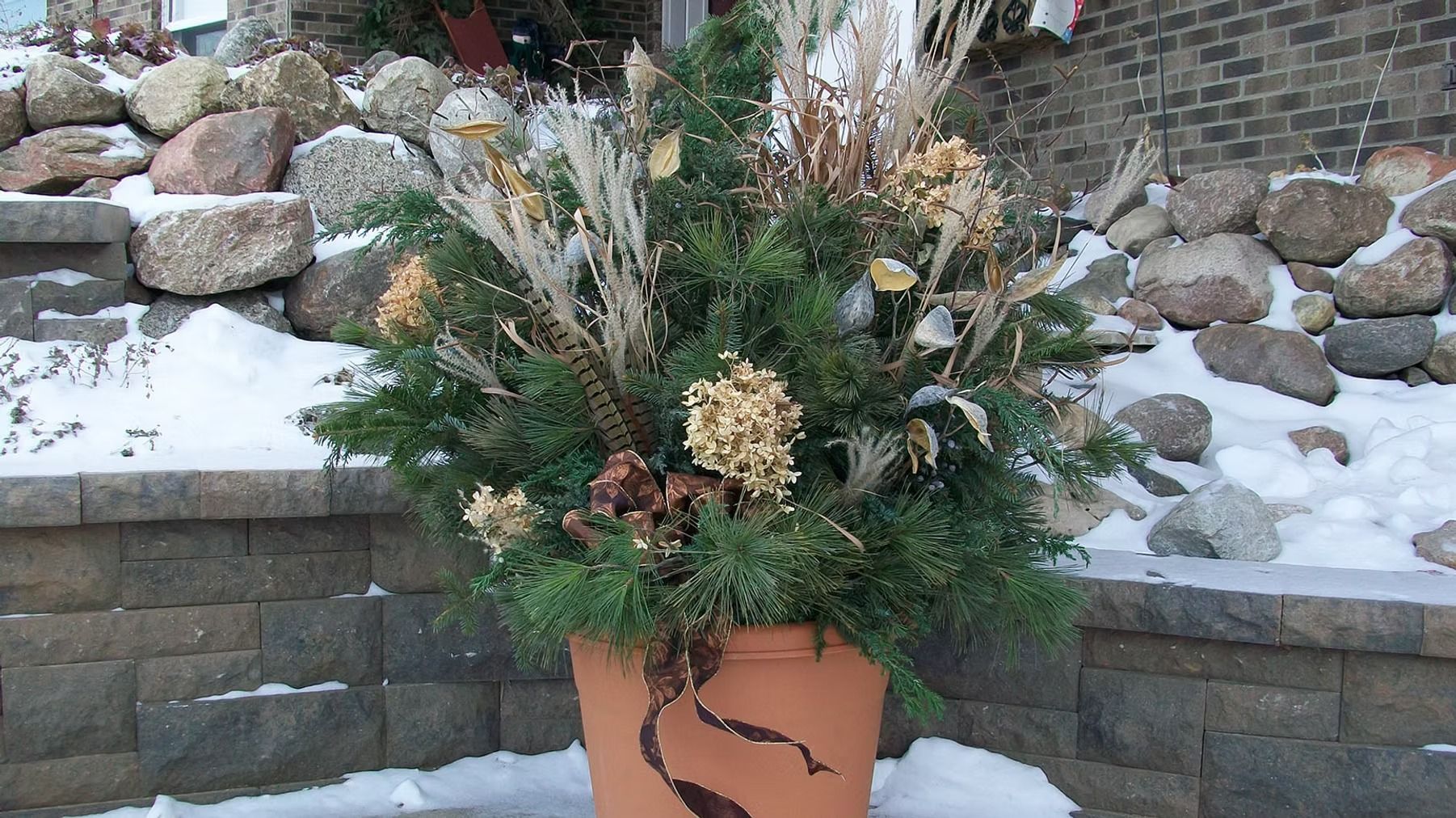 Winter planter with evergreen branches and dried flowers in a terracotta pot by a snowy stone wall