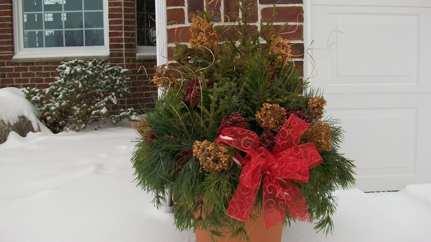 Snowy porch with a potted evergreen holiday arrangement topped with a large red bow.