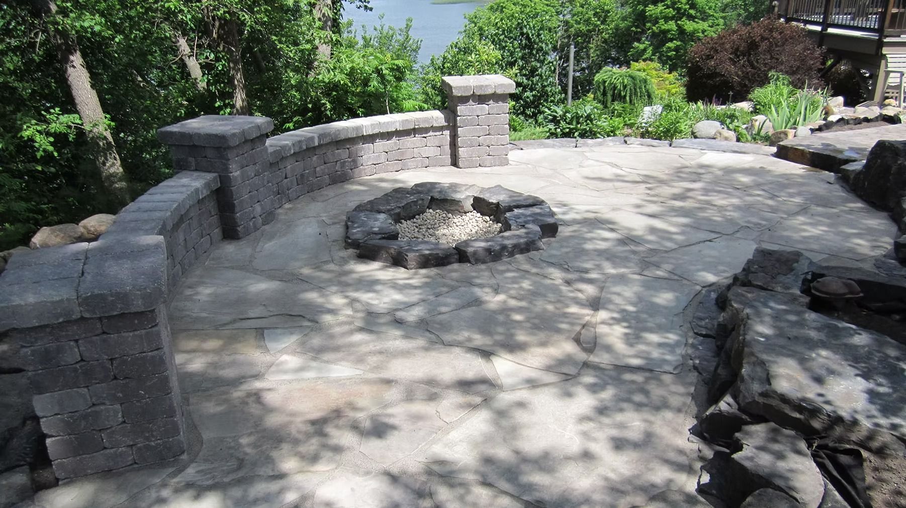 Stone patio with a circular fire pit, built-in seating wall, and surrounding greenery in a sunny backyard.