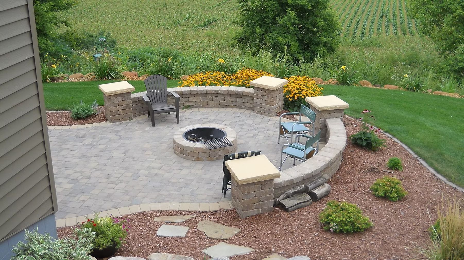 Backyard patio with circular stone fire pit, curved bench seating, and flower planters amid green lawn.