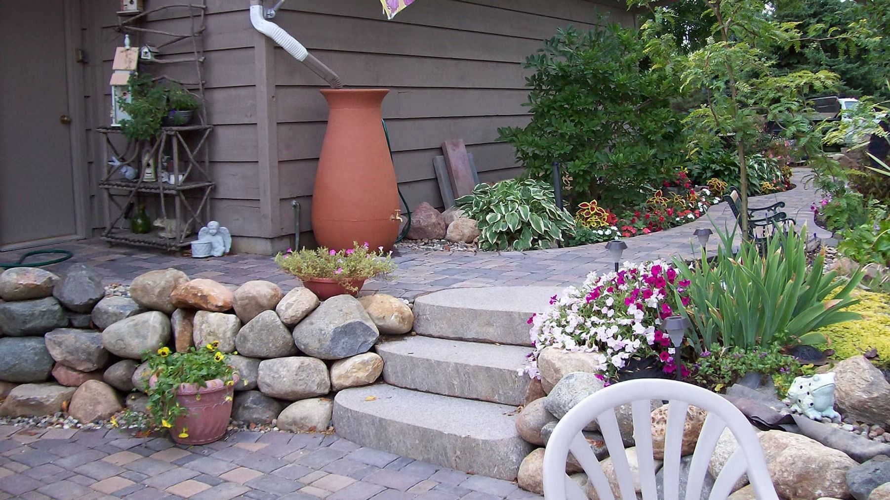 Front yard garden with stone steps, potted plants, terraced rocks, and flowers beside a house.