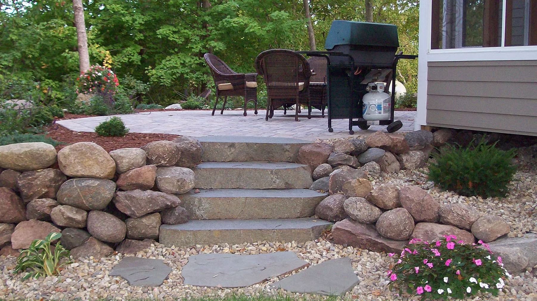 Stone steps leading to a patio with chairs, a grill, and greenery in a backyard