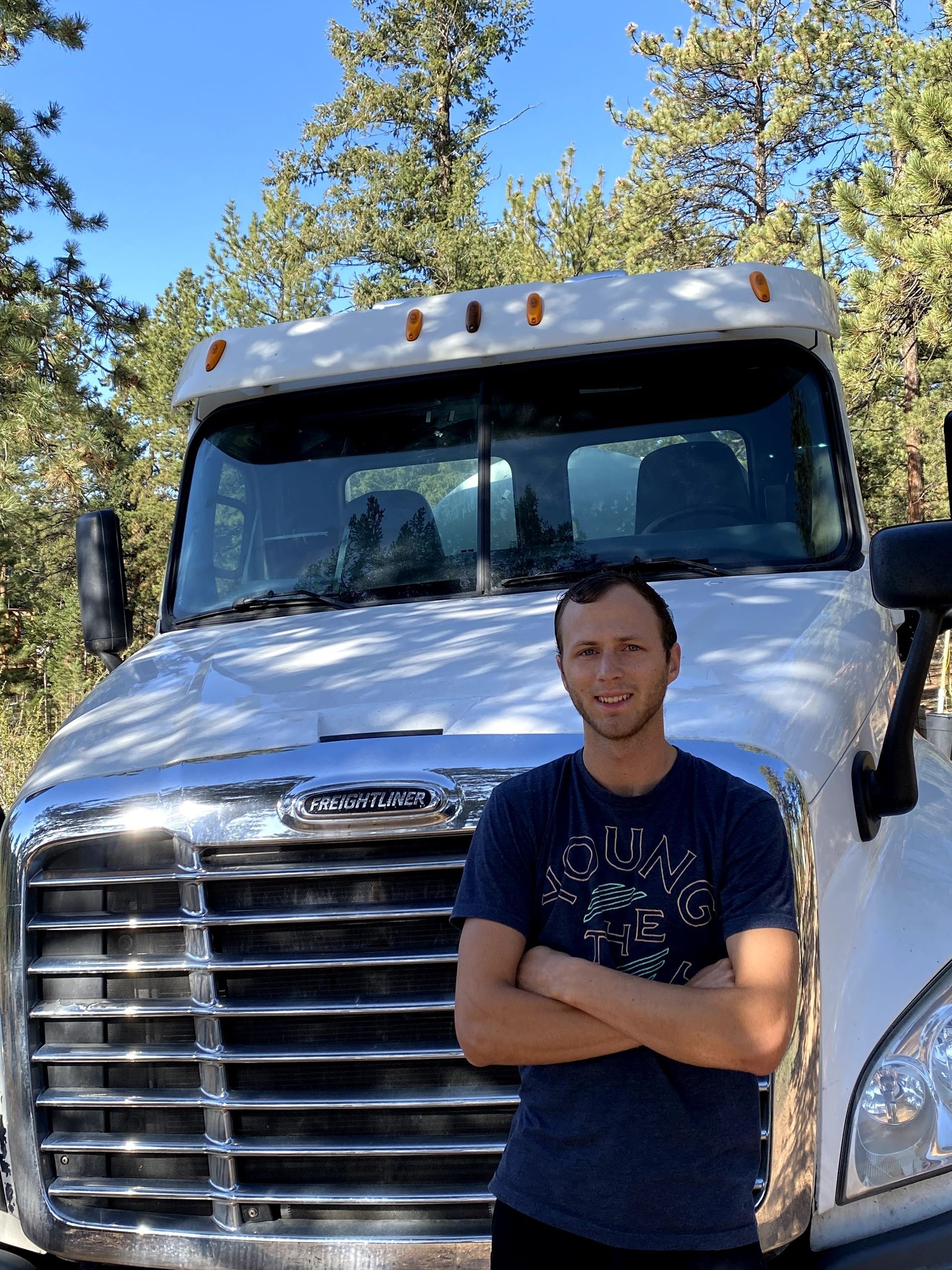 A man is standing in front of a semi truck with his arms crossed.