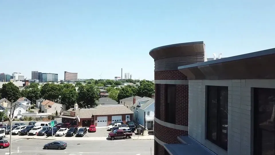 View from a building rooftop: cars on a street, other buildings, trees, and blue sky.