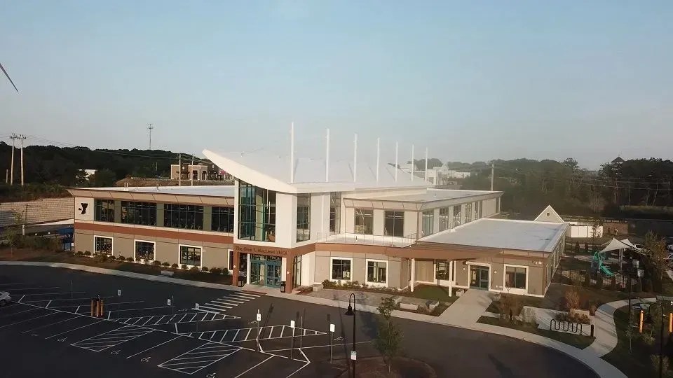 Modern two-story building with white roof and large windows, fronted by a parking lot.