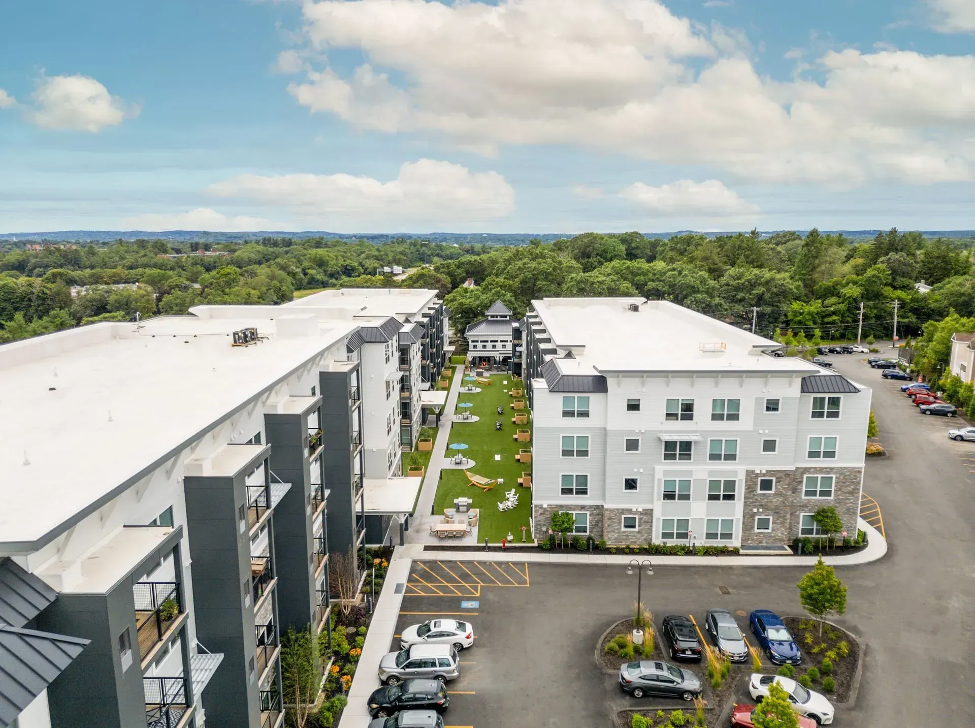 Aerial view of apartment complex with parking, green space, and surrounding trees under a cloudy sky.