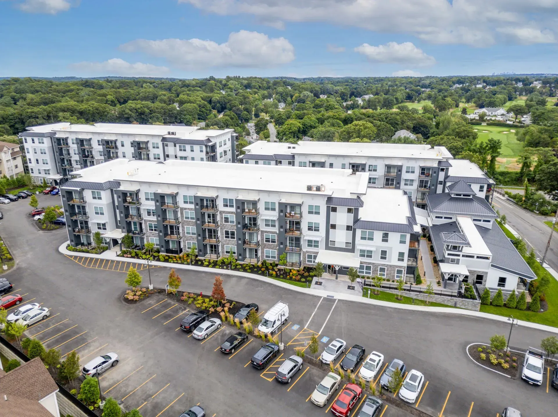 Aerial view of a multi-story apartment complex with parking lot, set against a backdrop of trees and a bright sky.
