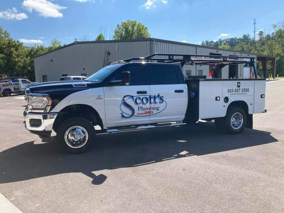White and blue plumbing service truck, "Scott's Plumbing," parked in front of a building on a sunny day.