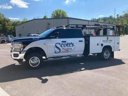 White and blue plumbing service truck, "Scott's Plumbing," parked in front of a building on a sunny day.