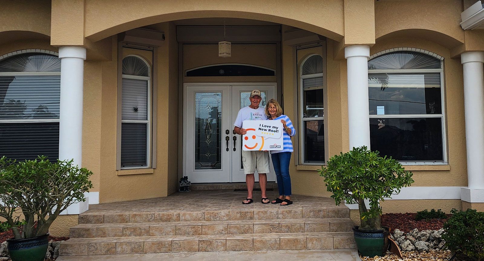 A man and woman are standing in front of a house holding a sign.