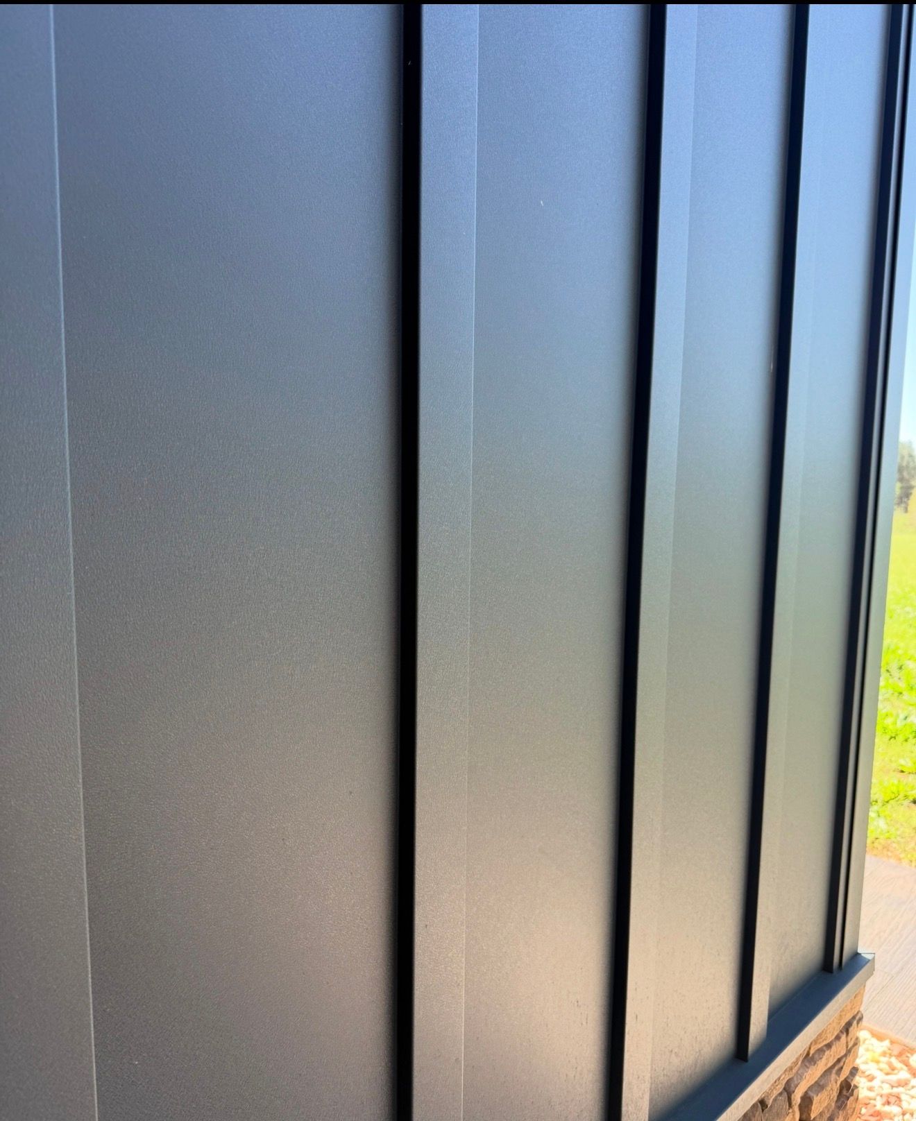 Close-up of a matte black metal wall or fence panel in sunlight with green grass visible at the edge