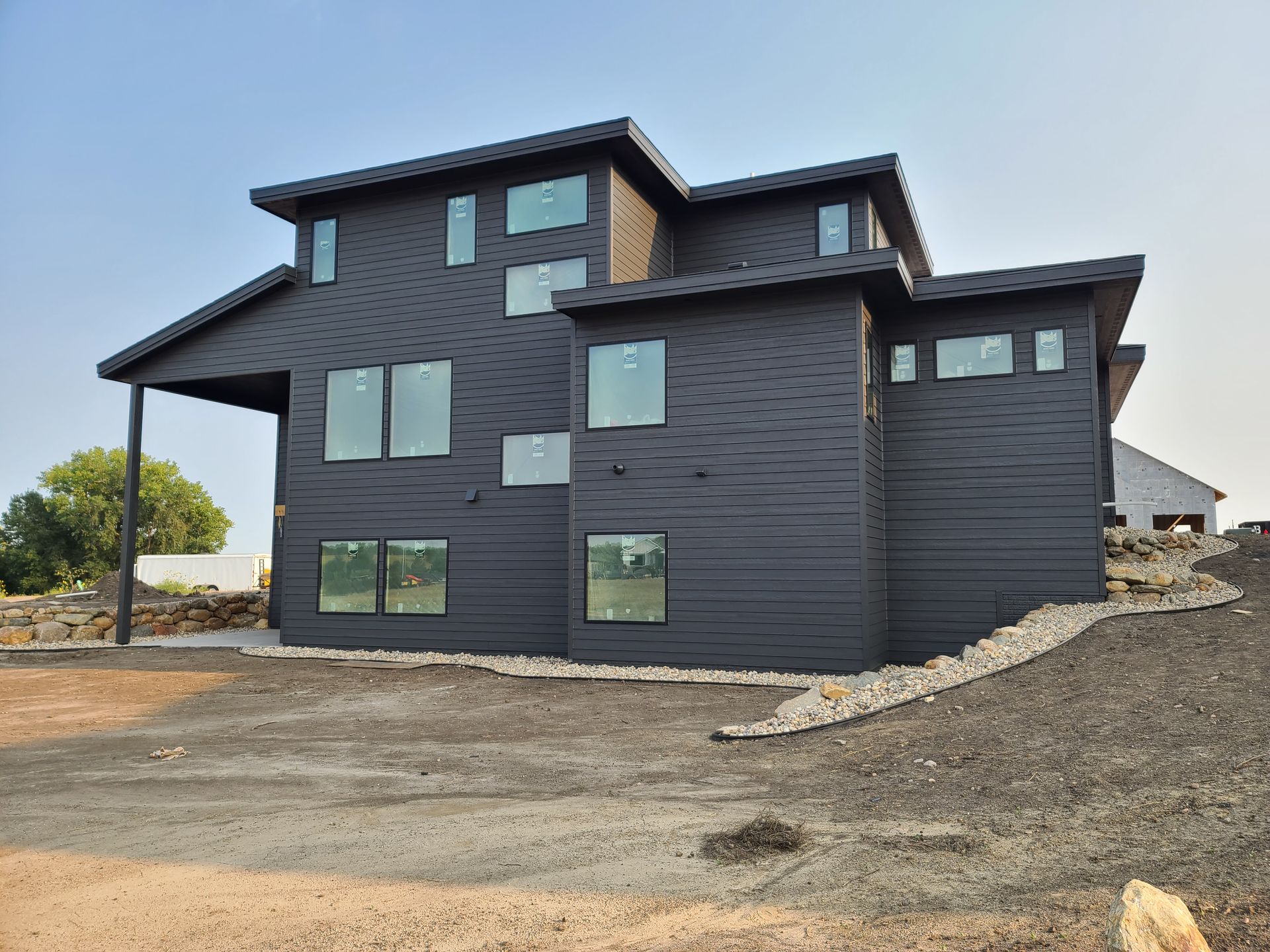 Modern, black-sided house with multiple rectangular windows, built on a slight hill, under a clear sky.