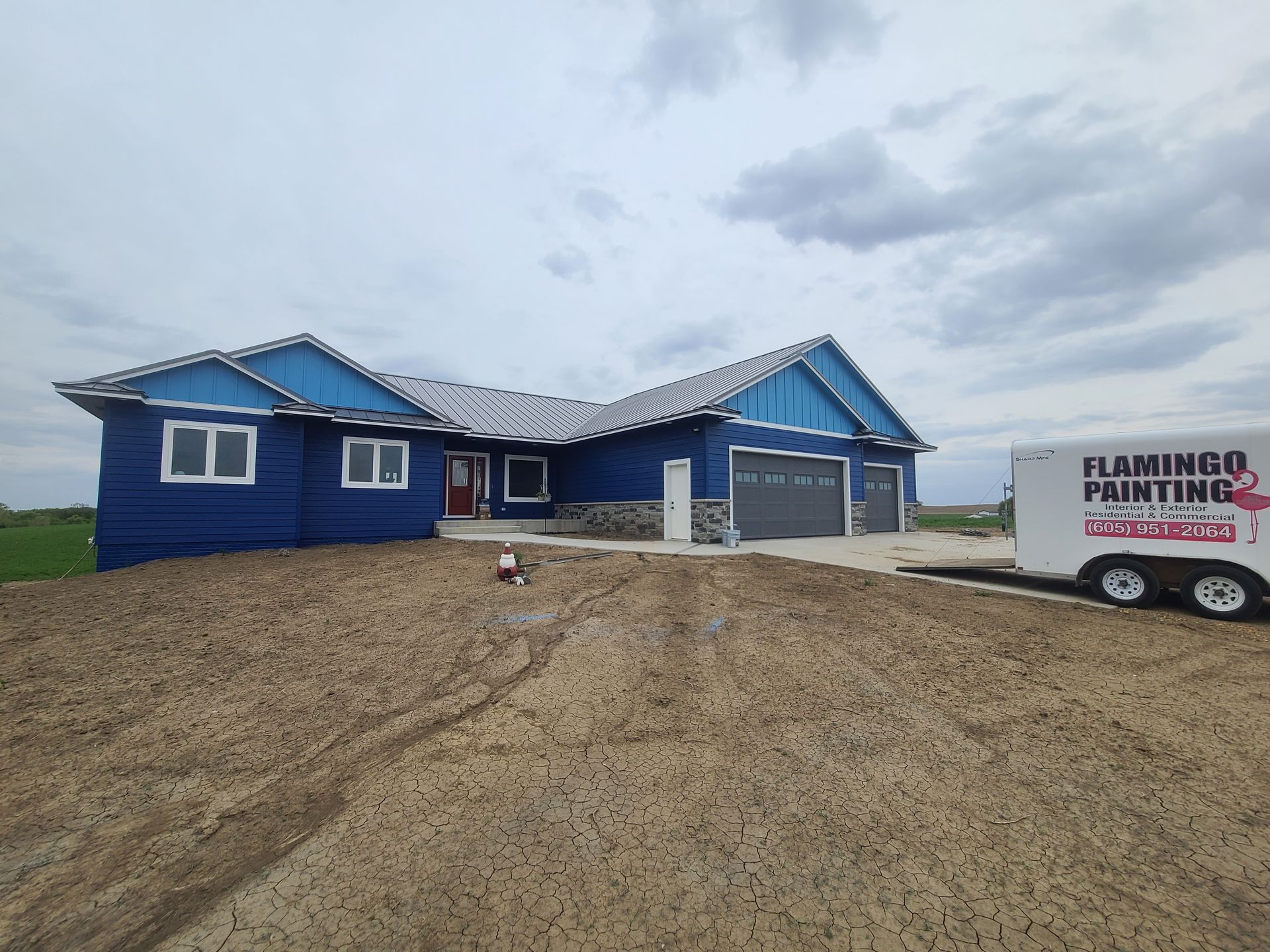 Blue house with grey garage doors, Flamingo Painting trailer nearby on dirt.
